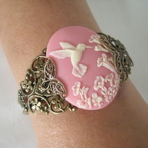 May include: A pink cameo bracelet with a white hummingbird and floral design. The bracelet is made of metal with a filigree design.