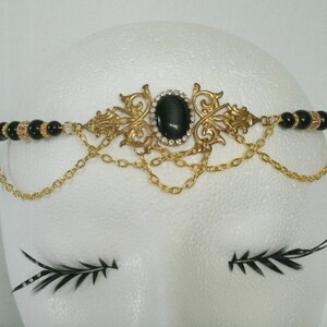 May include: A gold and black headband with a large black gemstone in the centre. The headband is adorned with intricate gold filigree and black beads.