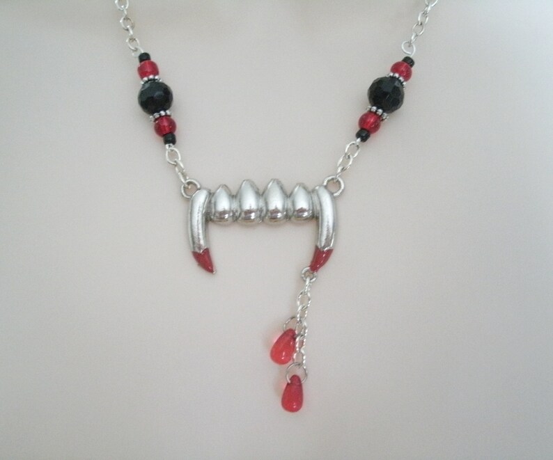 May include: Silver vampire fangs necklace with red and black beads. The necklace has a silver chain and a silver pendant shaped like a set of fangs with red tips. The pendant is attached to a silver chain with three red beads hanging from it.