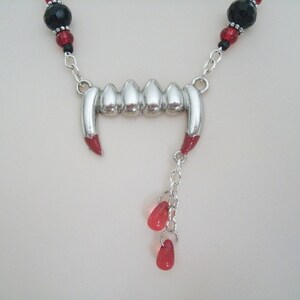 May include: A silver vampire fangs necklace with red and black beads. The necklace has a silver chain and a silver pendant with a row of fangs. The pendant is attached to a silver chain with three red beads.