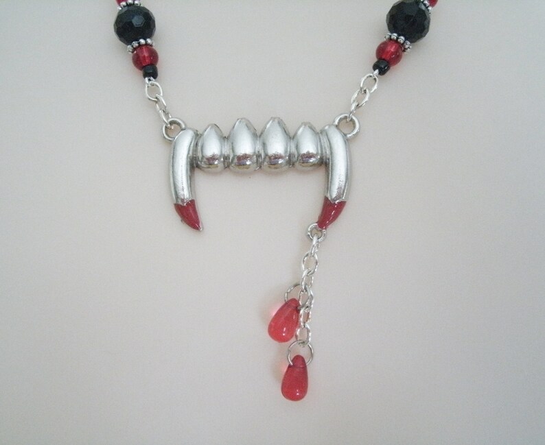 May include: A silver vampire fangs necklace with red and black beads. The necklace has a silver chain and a silver pendant with a row of fangs. The pendant is attached to a silver chain with three red beads.