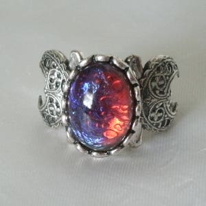 May include: A silver-toned ring with an oval, iridescent, red and purple stone. The ring has ornate, scroll-like details on the band and around the stone setting. The ring is a piece of jewelry.
