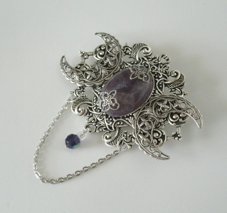 May include: A silver filigree brooch with a large purple gemstone in the center. The brooch has a chain attached to it with a small purple bead at the end.