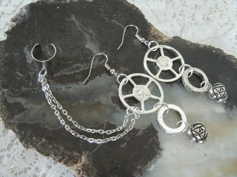 May include: A pair of silver earrings with a chain connecting them. Each earring features a circular design with a smaller circle dangling below. The smaller circle has a small ball attached to it.
