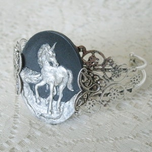 May include: A silver bracelet with a black cameo featuring a silver unicorn. The cameo is surrounded by a silver filigree design.