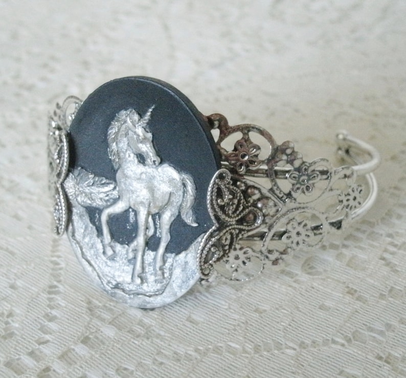May include: A silver bracelet with a black cameo featuring a silver unicorn. The cameo is surrounded by a silver filigree design.