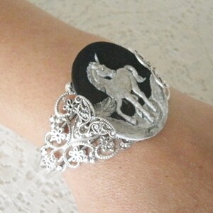 May include: A silver bracelet with a black cameo featuring a silver unicorn. The bracelet has a filigree design with paisley shapes and flowers.