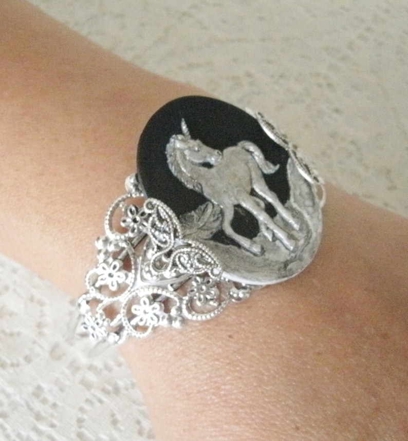 May include: A silver bracelet with a black cameo featuring a silver unicorn. The bracelet has a filigree design with paisley shapes and flowers.