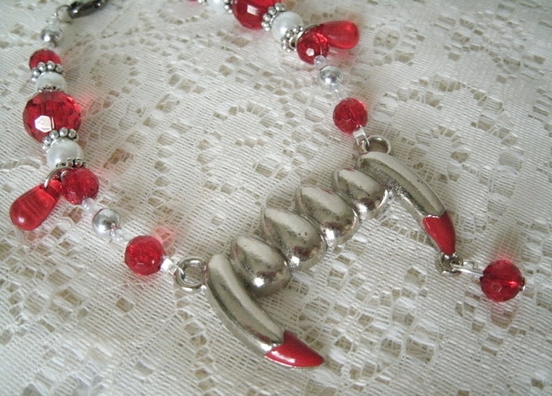 May include: A silver charm necklace with a fang pendant. The necklace is made with red and white beads.