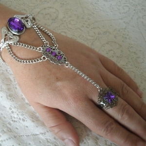 May include: A silver chain bracelet with a purple gemstone and a matching ring. The bracelet has a delicate design and is connected to the ring by a chain.