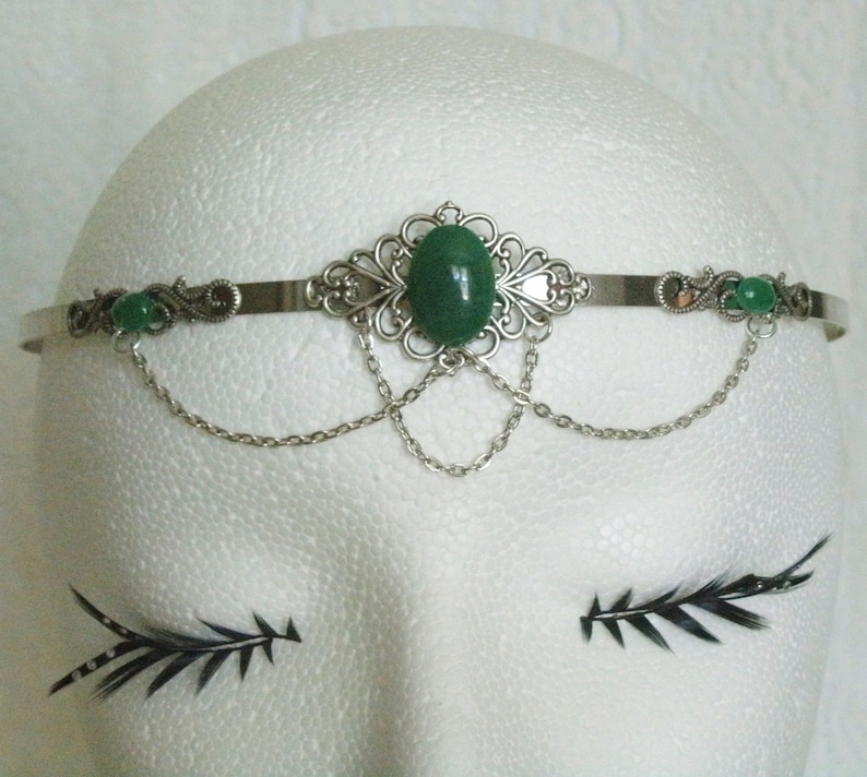 May include: Silver headband with a green gemstone and dangling chains. The headband is decorated with ornate silver details.