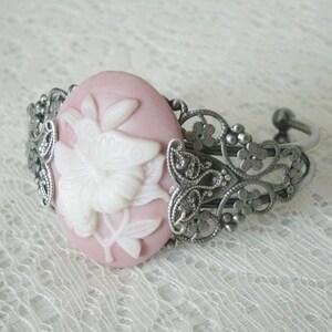 May include: A pink cameo bracelet with a white butterfly design. The bracelet is made of silver tone metal and has a delicate filigree design.