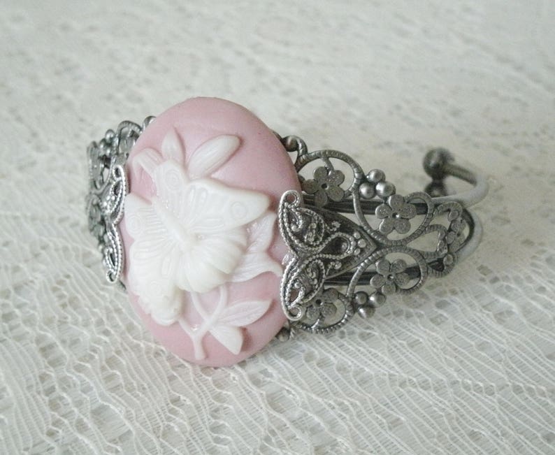 May include: A pink cameo bracelet with a white butterfly design. The bracelet is made of silver tone metal and has a delicate filigree design.