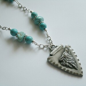 May include: A silver necklace with a turquoise and silver arrowhead pendant featuring a wolf's head. The arrowhead pendant has a textured surface.