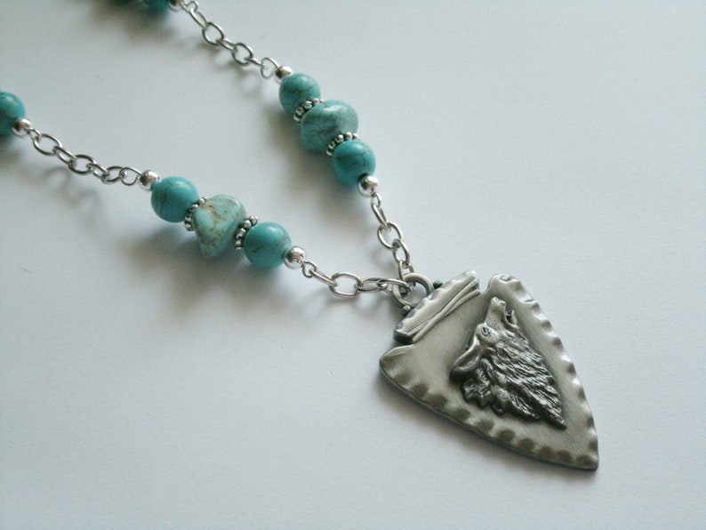 May include: A silver necklace with a turquoise and silver arrowhead pendant featuring a wolf's head. The arrowhead pendant has a textured surface.