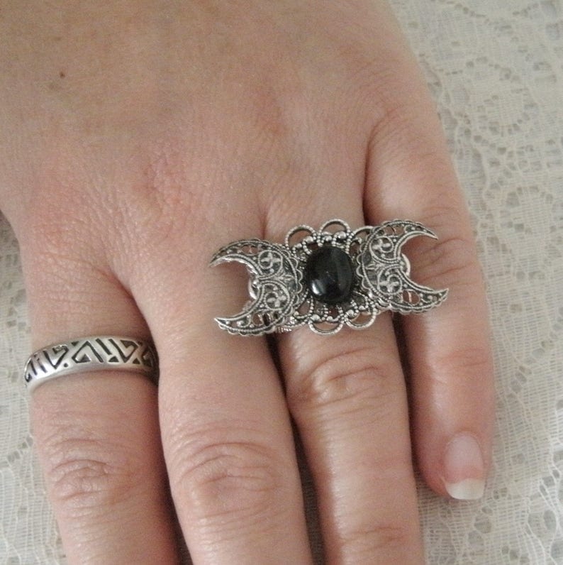 May include: A silver ring with a black stone and a triple moon design. The ring is on a finger.