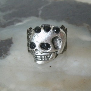 May include: A silver skull ring with black rhinestone eyes. The ring is detailed with a textured skull design.