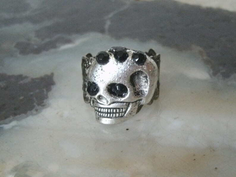 May include: A silver skull ring with black rhinestone eyes. The ring is detailed with a textured skull design.