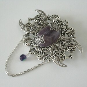 May include: A silver filigree brooch with a purple stone in the center and a dangling chain with a purple bead. The brooch features a crescent moon design.