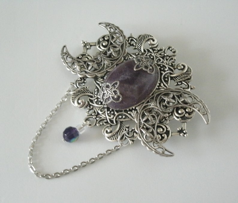 May include: A silver filigree brooch with a purple stone in the center and a dangling chain with a purple bead. The brooch features a crescent moon design.