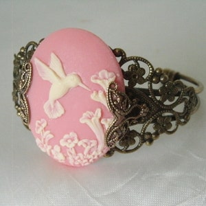 May include: A pink cameo bracelet with a white hummingbird and floral design. The bracelet is made of metal with a filigree pattern.