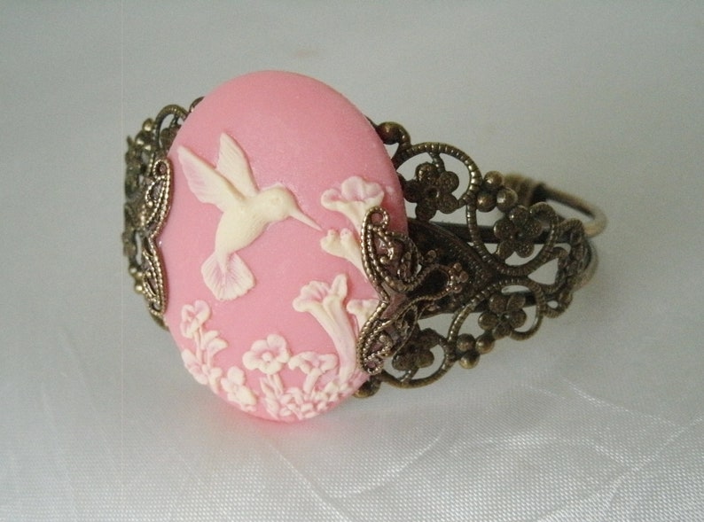 May include: A pink cameo bracelet with a white hummingbird and floral design. The bracelet is made of metal with a filigree pattern.