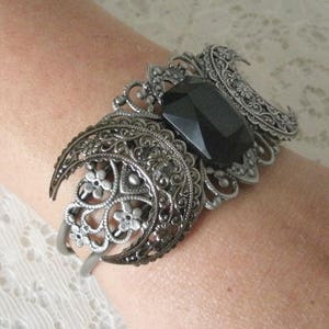 May include: Silver bracelet with a large black gemstone in the center, surrounded by intricate filigree designs. The bracelet features two crescent moon shapes on either side of the gemstone.