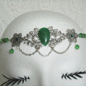 Puede incluir: Diadema de metal plateado y verde con una piedra verde en forma de l&aacute;grima en el centro. La diadema est&aacute; decorada con dise&ntilde;os florales y tiene un detalle de cadena.