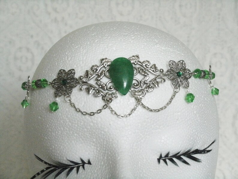 Puede incluir: Diadema de metal plateado y verde con una piedra verde en forma de l&aacute;grima en el centro. La diadema est&aacute; decorada con dise&ntilde;os florales y tiene un detalle de cadena.