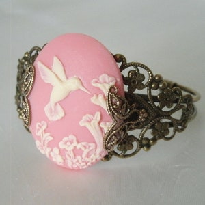 May include: A pink cameo bracelet with a white hummingbird and floral design. The bracelet is made of metal with a filigree design.