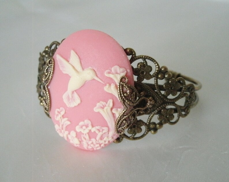 May include: A pink cameo bracelet with a white hummingbird and floral design. The bracelet is made of metal with a filigree design.