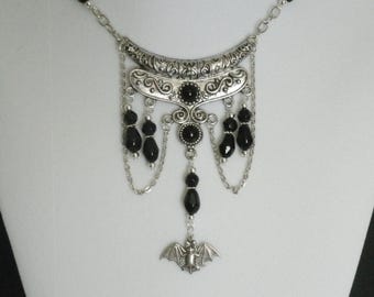 Puede incluir: Un collar plateado y negro con un colgante de murciélago. El collar presenta una pieza central de plata grande y adornada con cuentas negras y cadenas.