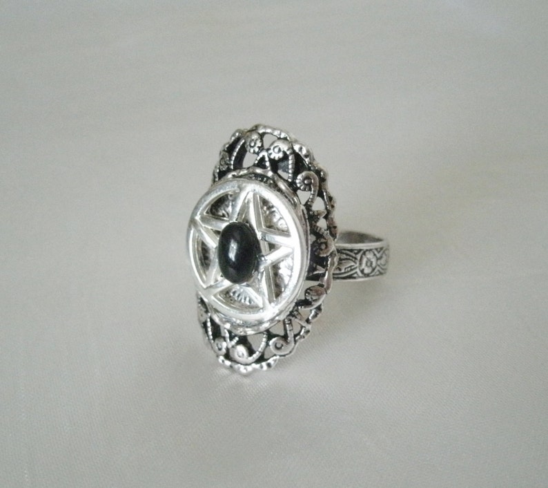 Puede incluir: Un anillo de plata con una piedra negra engastada en un dise&ntilde;o de pentagrama. El anillo tiene un dise&ntilde;o delicado y ornamentado.
