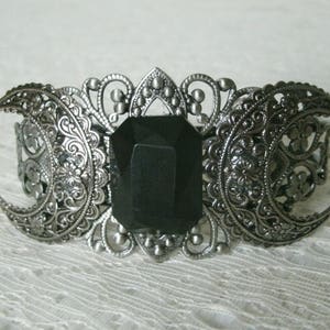 May include: A silver-tone bracelet with a black gemstone in the center. The bracelet is decorated with three crescent moons and intricate filigree details.