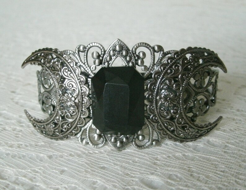 May include: A silver-tone bracelet with a black gemstone in the center. The bracelet is decorated with three crescent moons and intricate filigree details.