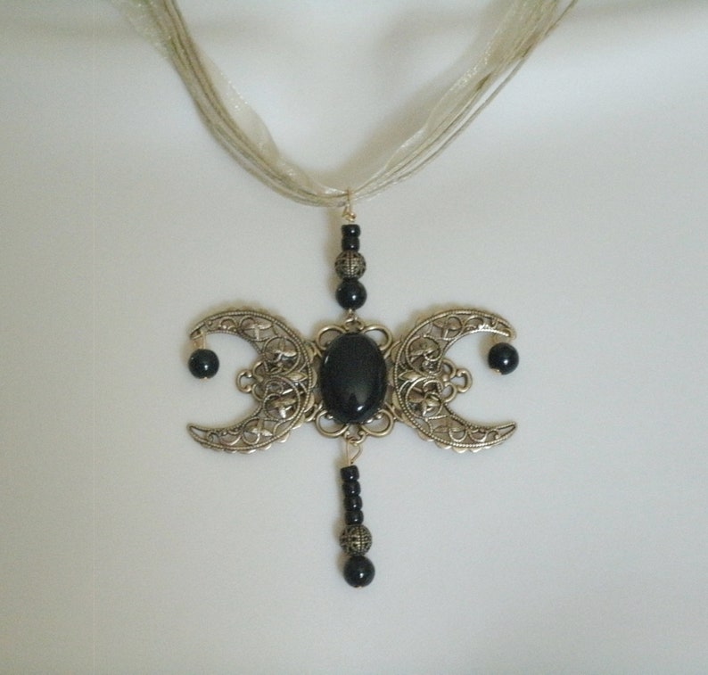 Puede incluir: Un collar con colgante plateado con un dise&ntilde;o de triple luna, un centro de piedra negra ovalada y detalles de cuentas negras. El colgante cuelga de una cinta plateada transparente.