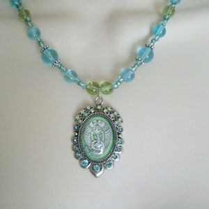 May include: A beaded necklace featuring a pendant with a light green oval center. The pendant has a silver-colored frame with blue and green accents. The necklace has blue and green beads.