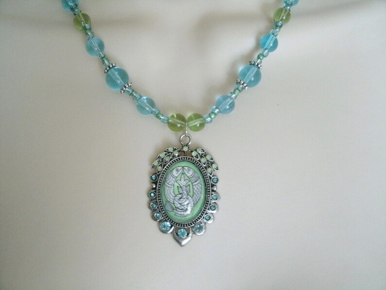 May include: A beaded necklace featuring a pendant with a light green oval center. The pendant has a silver-colored frame with blue and green accents. The necklace has blue and green beads.