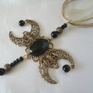 Puede incluir: Un collar artesanal con una piedra ovalada negra central, acentuado por formas de media luna de color bronce. Cuentas negras y una cadena de color bronce completan el dise&ntilde;o, suspendido de cintas beige.