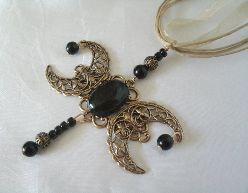 Puede incluir: Un collar artesanal con una piedra ovalada negra central, acentuado por formas de media luna de color bronce. Cuentas negras y una cadena de color bronce completan el dise&ntilde;o, suspendido de cintas beige.