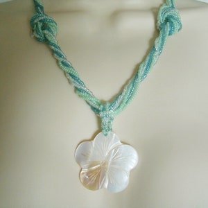 May include: A twisted necklace made of light blue and green seed beads with a large white shell flower pendant.