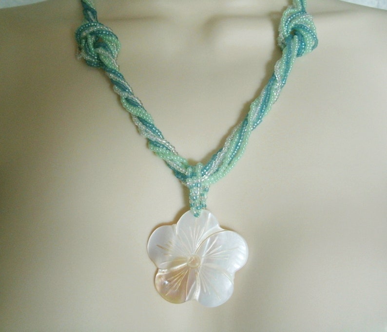 May include: A twisted necklace made of light blue and green seed beads with a large white shell flower pendant.