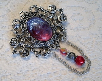 May include: A silver filigree brooch with a large, iridescent, pink and blue glass stone. The brooch has a chain with a red glass bead and a silver bead.
