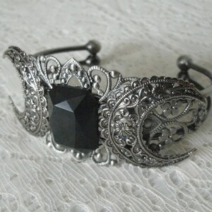May include: A silver bracelet with a black gemstone in the center and a filigree design. The bracelet is shaped like two crescent moons.