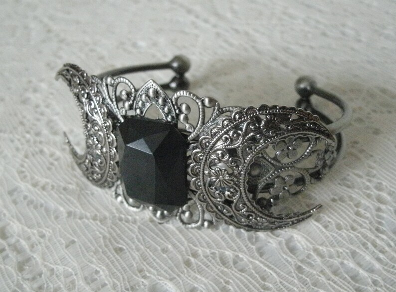 May include: A silver bracelet with a black gemstone in the center and a filigree design. The bracelet is shaped like two crescent moons.