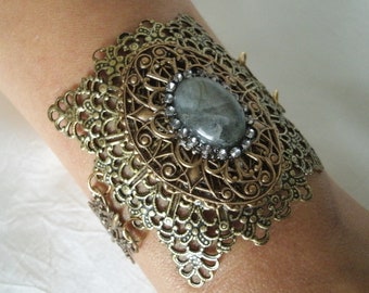 May include: A gold-toned metal bracelet with a large, oval-shaped gray gemstone set in the center. The bracelet is intricately designed with a filigree pattern and features small, clear rhinestones around the gemstone.