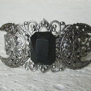 May include: A silver-tone bracelet with a black gemstone in the center. The bracelet is decorated with intricate filigree designs and three crescent moons.