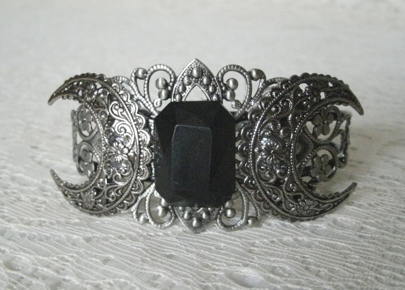 May include: A silver-tone bracelet with a black gemstone in the center. The bracelet is decorated with intricate filigree designs and three crescent moons.