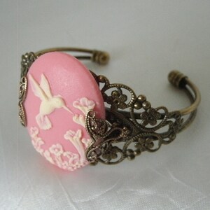 May include: A pink cameo bracelet with a delicate filigree design. The cameo features a white hummingbird in flight with pink flowers.