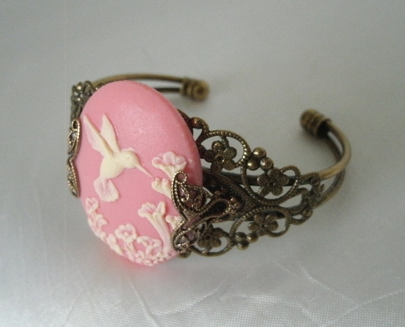 May include: A pink cameo bracelet with a delicate filigree design. The cameo features a white hummingbird in flight with pink flowers.